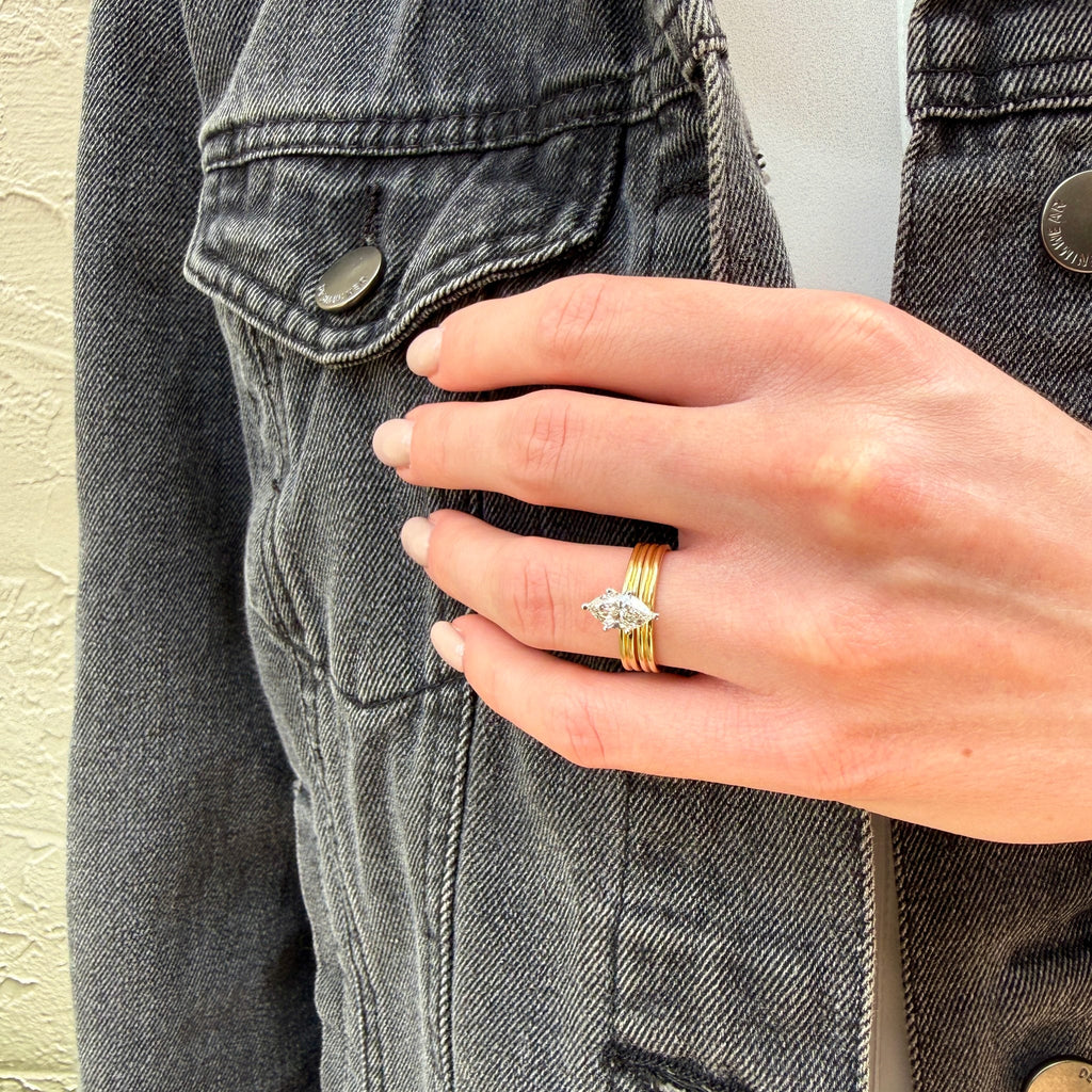 A close-up of a hand wearing a delicate gold ring with a small silver-toned leaf or feather-shaped stone, styled on top of a dark gray denim jacket.