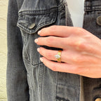 A close-up of a hand wearing a delicate gold ring with a small silver-toned leaf or feather-shaped stone, styled on top of a dark gray denim jacket.