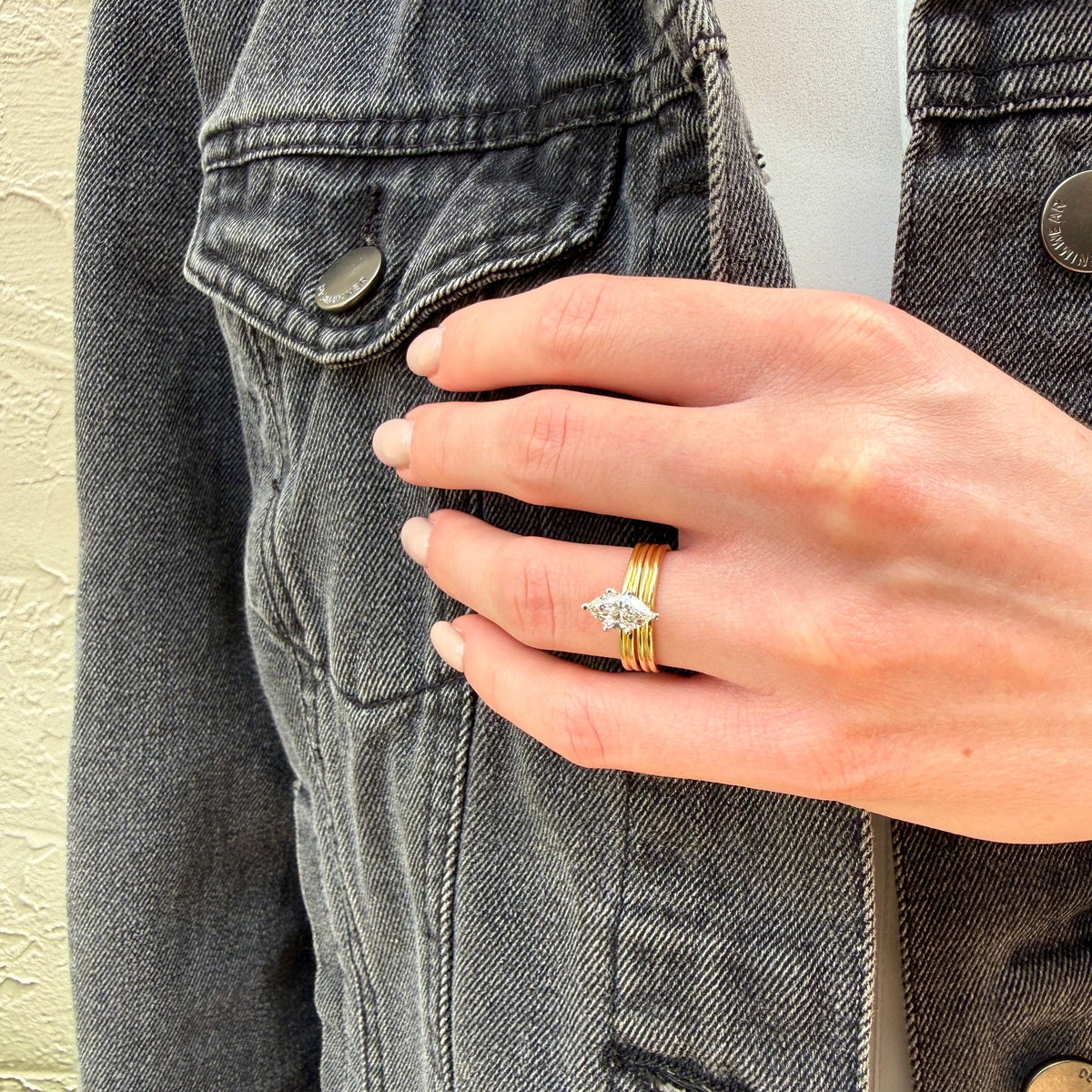 A close-up of a hand wearing a delicate gold ring with a small silver-toned leaf or feather-shaped stone, styled on top of a dark gray denim jacket.