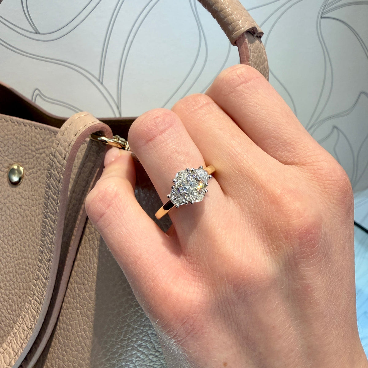 Hand wearing a diamond ring with a beige handbag in the background