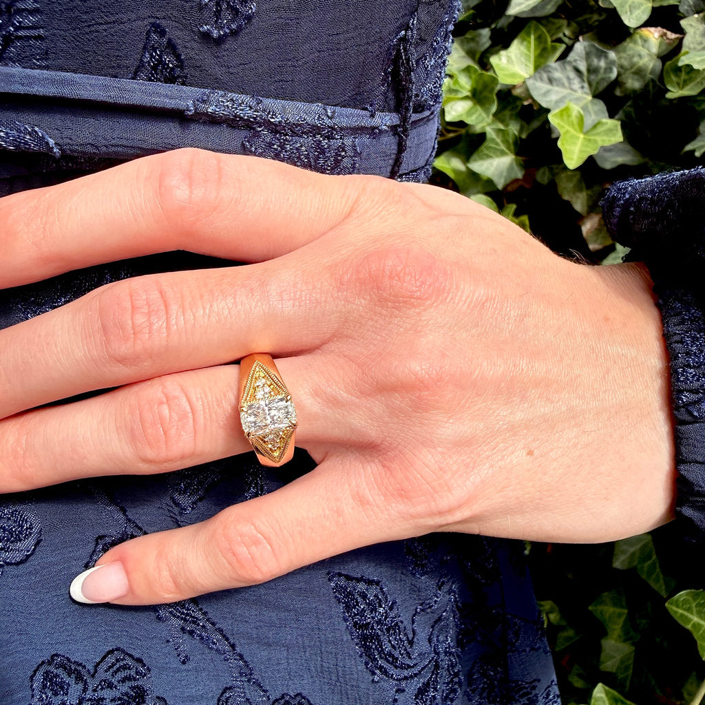 A close-up of a hand wearing a yellow gold ring with a marquise-shaped diamond setting, resting on a dark navy blue patterned dress with green ivy in the background.