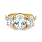 A yellow gold three-stone ring featuring a large oval center diamond flanked by two smaller oval side diamonds, with small accent diamonds along the shoulders, set against a plain white background.
