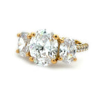 A yellow gold three-stone diamond ring featuring a large oval center diamond flanked by two smaller round diamonds, with a pavé-set diamond band, shown on a white background.