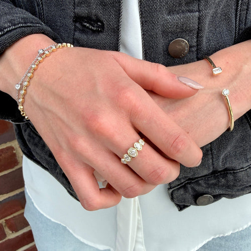 A close-up of a person's hands wearing delicate jewelry, including a gold ring with three round clear stones, a gold cuff bracelet with a rectangular clear stone, and a gold beaded bracelet with clear stones, styled with a white shirt and dark denim jacket.