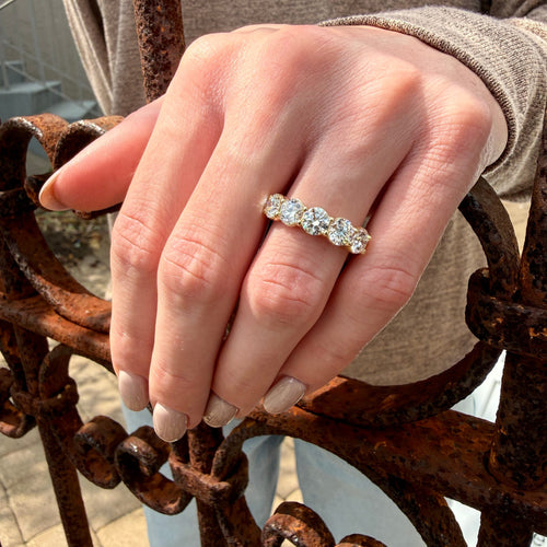 A close-up of a hand wearing a five-stone diamond ring with a gold band, resting on a rusty wrought iron fence.