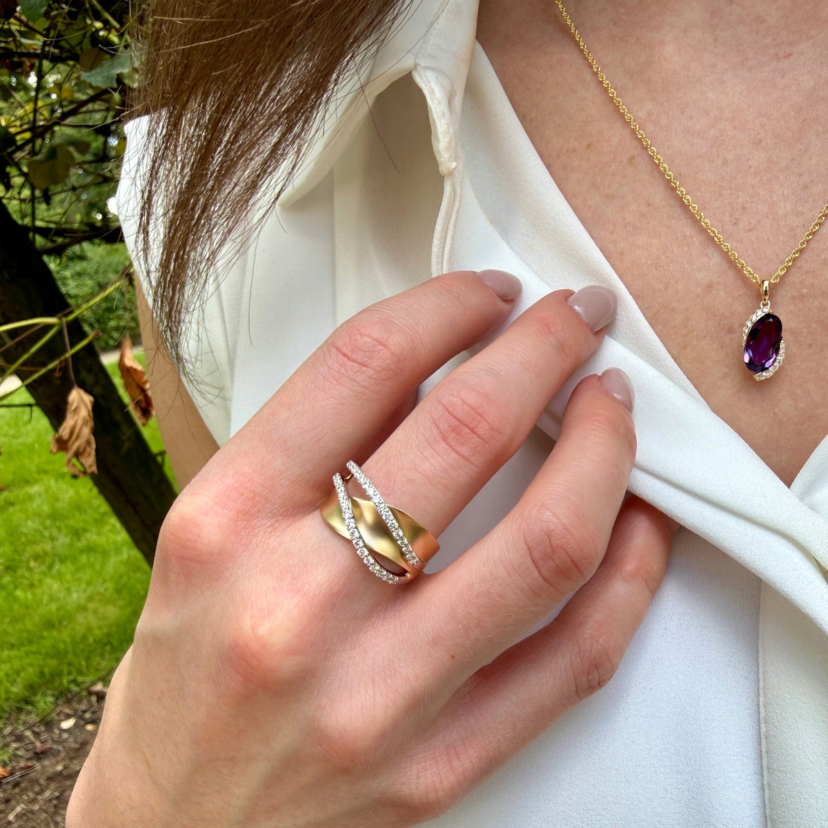 A close-up lifestyle shot of a person's hand wearing a double-band ring with one polished gold band, one silver band set with small clear stones, and a white blouse. A gold necklace with a purple marquise gemstone is also visible against an outdoor background.