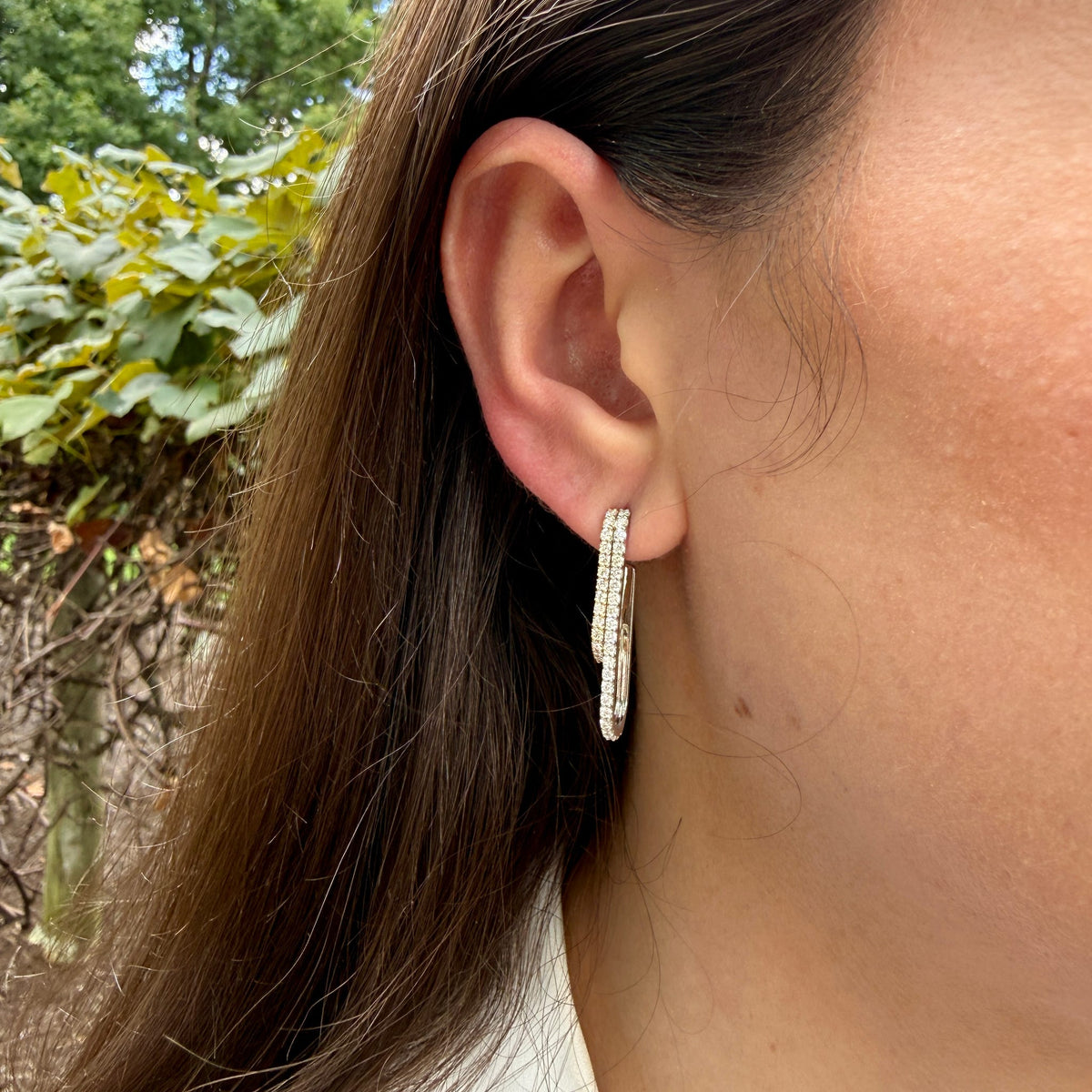 A close-up of a person's ear wearing a silver-toned huggie hoop earring paved with small clear stones, with outdoor greenery blurred in the background.