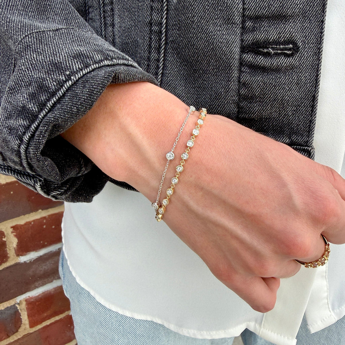 A close-up of a person's wrist wearing two delicate bracelets: one silver chain with clear stones and one gold chain with small gold beads and a central gold stone, styled with a white shirt and dark denim jacket.