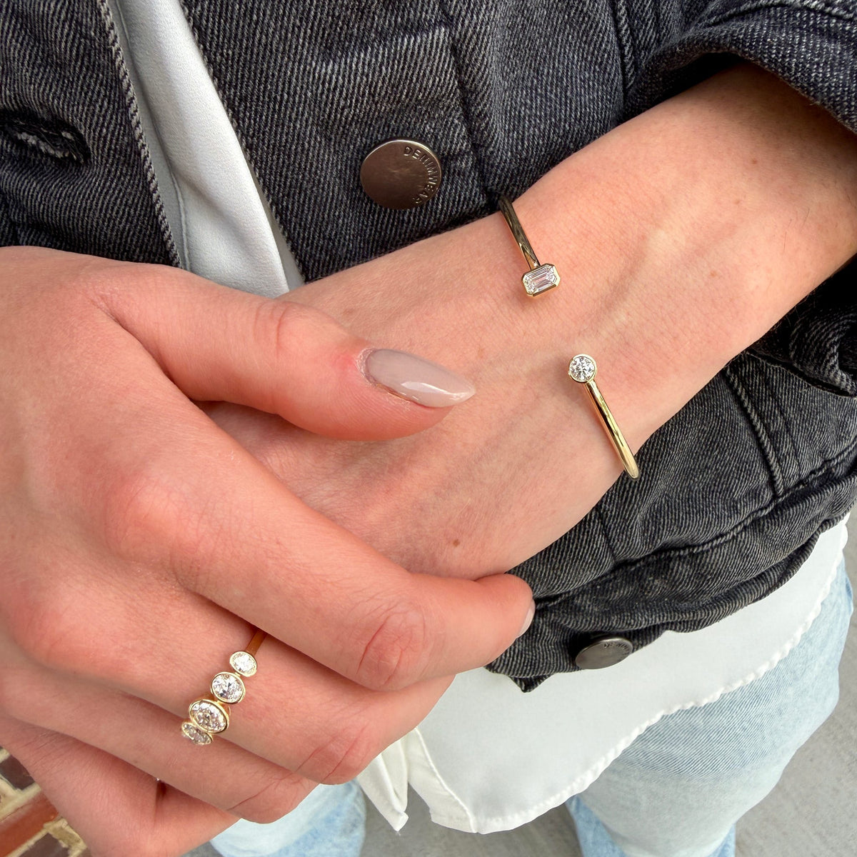A close-up of a person's hands and wrist wearing three delicate gold rings with clear gemstones, including a small band with a rectangular clear stone and two slim bands with round clear stones.