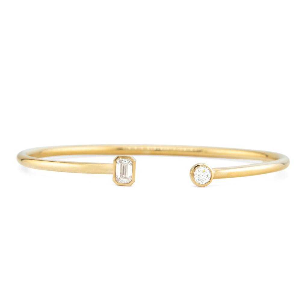 A delicate gold bangle bracelet with an open-ended design, featuring one rectangular emerald-cut clear stone and one round clear stone on a plain white background.