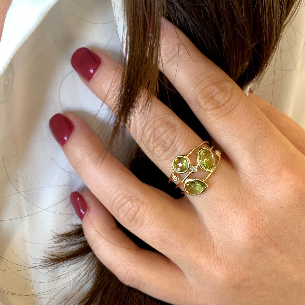 A close-up of a hand wearing a gold ring with three oval green gemstones, resting against dark brown hair and a white shirt.