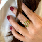 A close-up of a hand wearing a gold ring with three oval green gemstones, resting against dark brown hair and a white shirt.