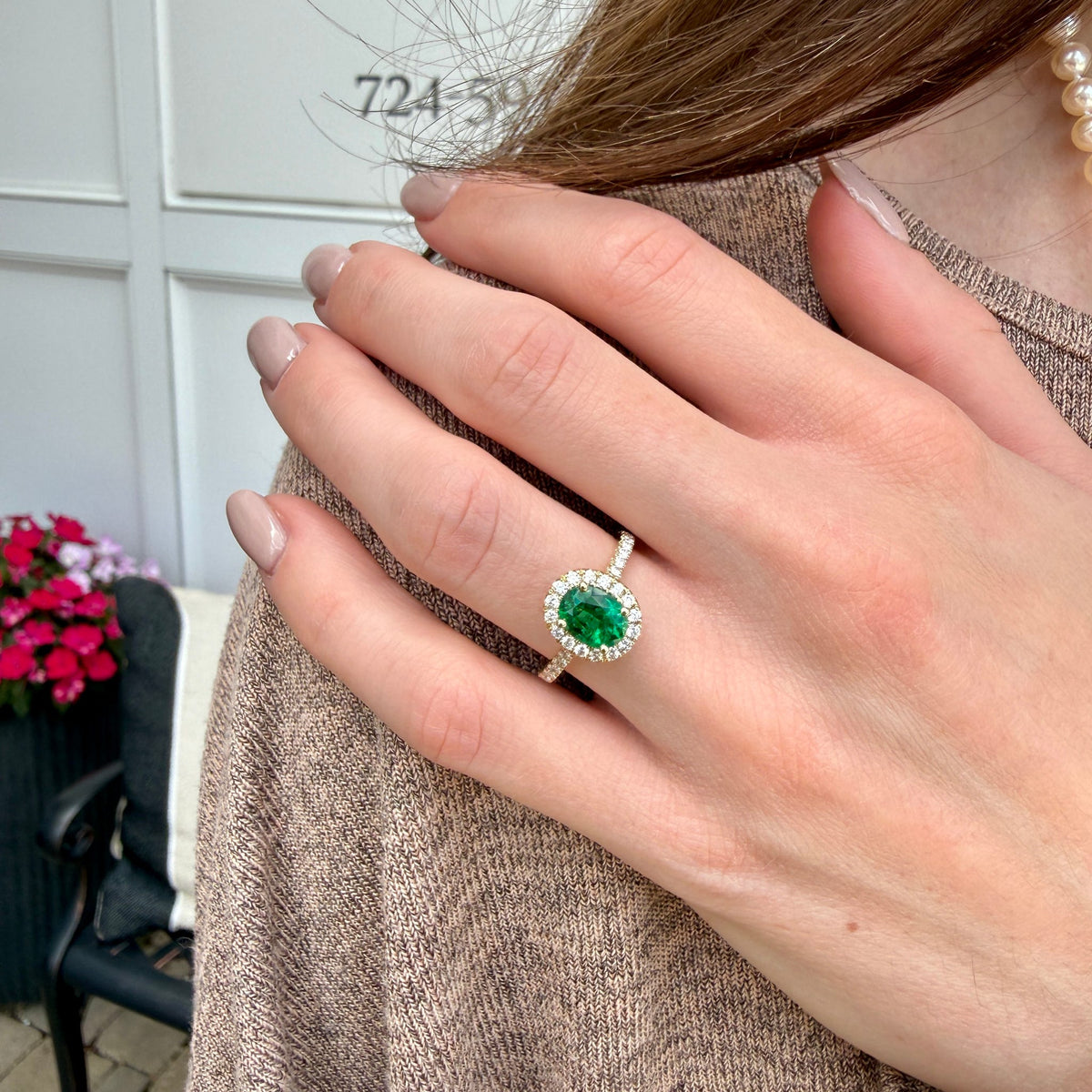 A close-up of a person's hand wearing a silver ring with a round green gemstone surrounded by a halo of small clear stones, set against a neutral indoor background.