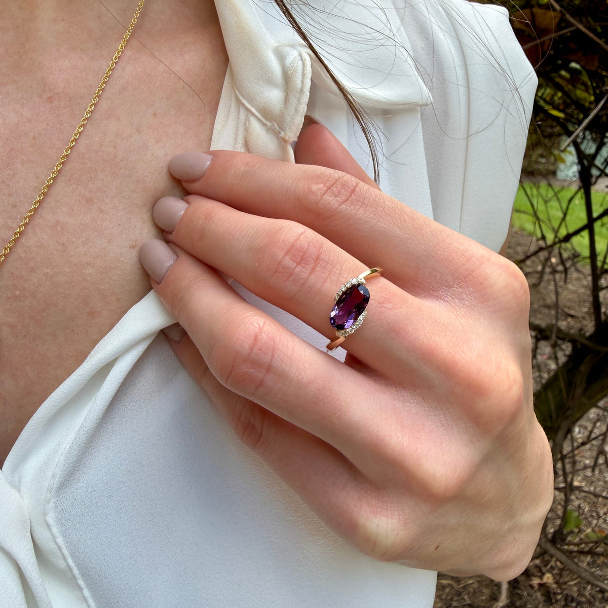 A close-up lifestyle shot of a woman wearing a rose gold ring with an oval purple gemstone, styled with a white blouse and a gold chain necklace.