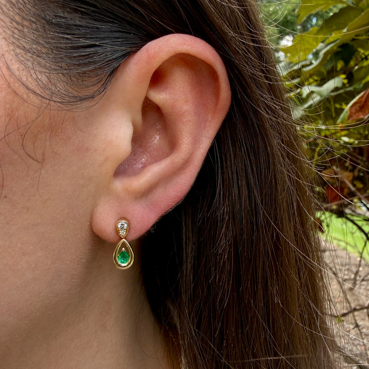 A close-up of a person's ear wearing a gold teardrop dangle earring with a green teardrop gemstone and a small clear stone above it, set against an outdoor background with green foliage.