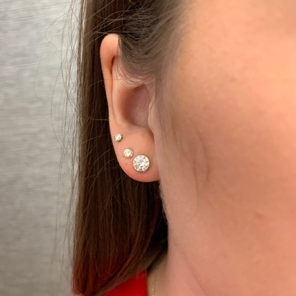 A close-up side view of a person's ear wearing three round stud earrings in silver-toned settings. The largest earring is on the lobe, with two smaller studs above it.