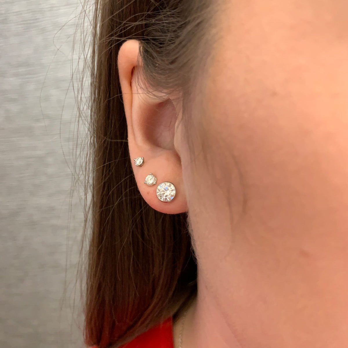 A close-up side view of a person's ear wearing three round stud earrings in silver-toned settings. The largest earring is on the lobe, with two smaller studs above it.