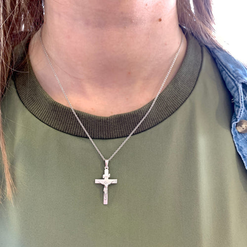 A silver crucifix pendant necklace worn by a person in an olive green shirt, with a denim jacket visible on the side.