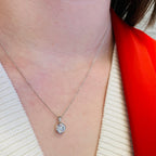 A close-up of a person's neck and chest wearing a delicate silver chain necklace with a round bezel-set clear gemstone pendant, styled with a cream knit top and a bright red fabric accent.