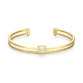 A gold-plated open cuff bracelet featuring a central rectangular bezel-set clear gemstone, with one side of the band wrapped in a textured braid pattern and the other side in smooth polished metal.