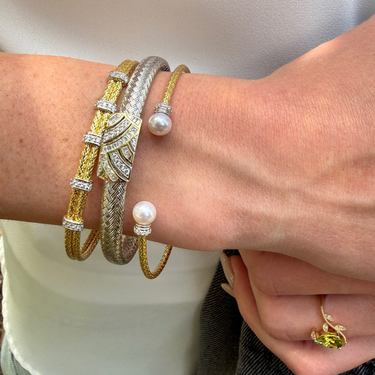 A close-up of a person's wrist wearing four stacked bracelets: two gold and two silver, including textured bands, pavé-set stones, and pearl accents.