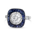 A vintage-style square ring featuring a large round brilliant-cut diamond in the center, surrounded by a halo of deep blue sapphires, with a silver-toned band accented by small side diamonds.