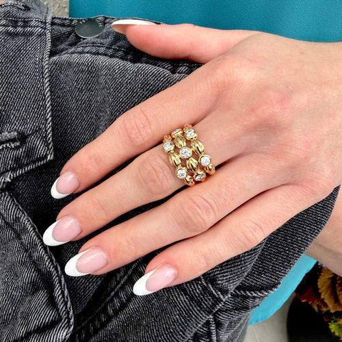 A close-up of a hand wearing a gold ring with multiple rows of round clear gemstones, resting on dark gray denim jeans.