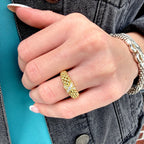 A close-up of a person's hand wearing a textured gold ring with a small clear center stone, paired with a silver chain bracelet, set against a denim jacket and teal shirt background.