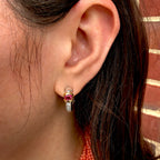 A close-up of a person's ear wearing a small gold huggie hoop earring with a pinkish-red rectangular center stone and a clear stone below it, against a brick wall background.
