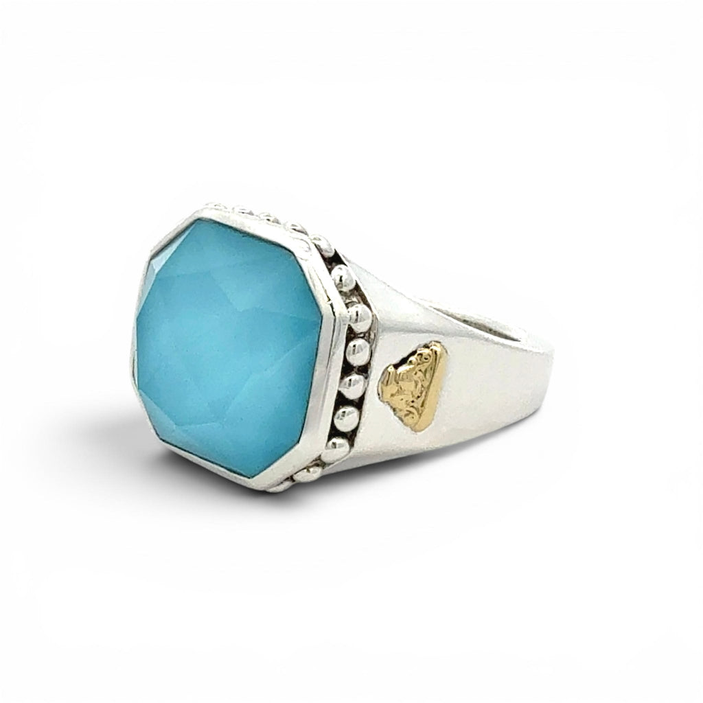 A silver-toned octagonal ring featuring a faceted light blue gemstone, accented with small silver beads on one side and a small gold heart-shaped emblem on the other, set against a plain white background.