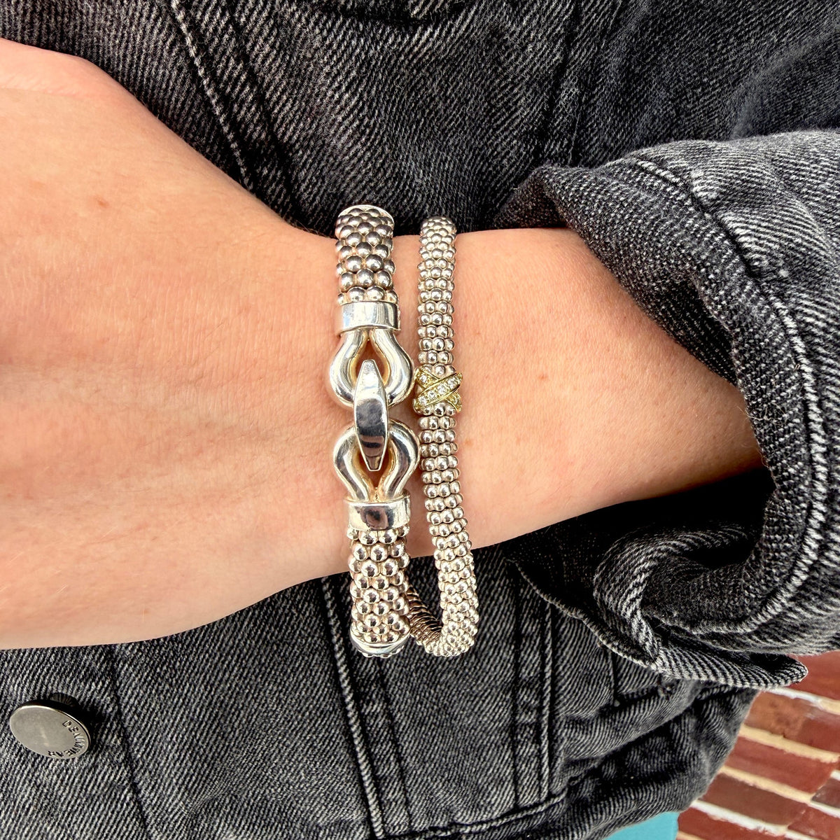 Two silver-toned bracelets worn on a person's wrist, including a textured chain link bracelet and a beaded bracelet with a small gold accent, set against a dark gray denim jacket.