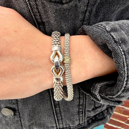 Two silver-toned bracelets worn on a person's wrist, including a textured chain link bracelet and a beaded bracelet with a small gold accent, set against a dark gray denim jacket.