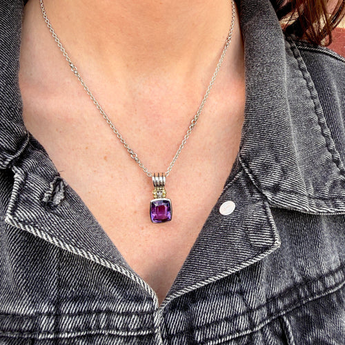 A close-up of a person wearing a silver chain necklace with a rectangular purple gemstone pendant, styled with a dark gray denim jacket.