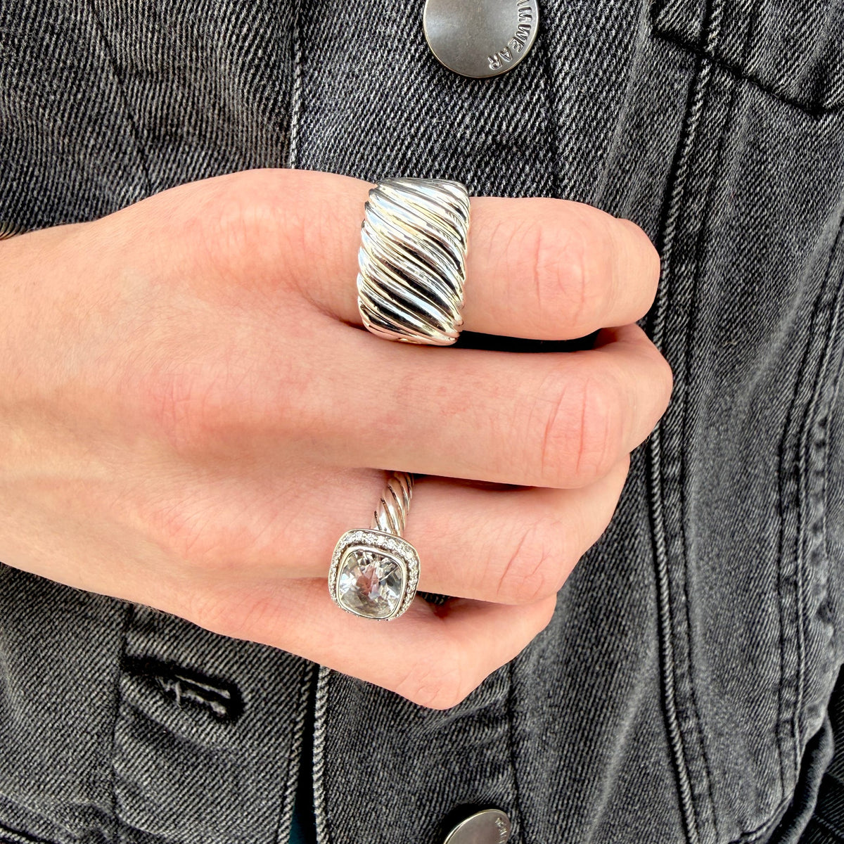 A close-up of a hand wearing two silver rings against a dark gray denim jacket. The larger ring on the middle finger has a twisted, ribbed band, while the smaller ring on the ring finger features a square clear gemstone in a silver setting.