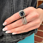 A close-up of a hand wearing a silver-toned ring with a square-cut dark blue gemstone, surrounded by a halo setting and a matching multi-band ring, against a denim jacket and brick wall background.
