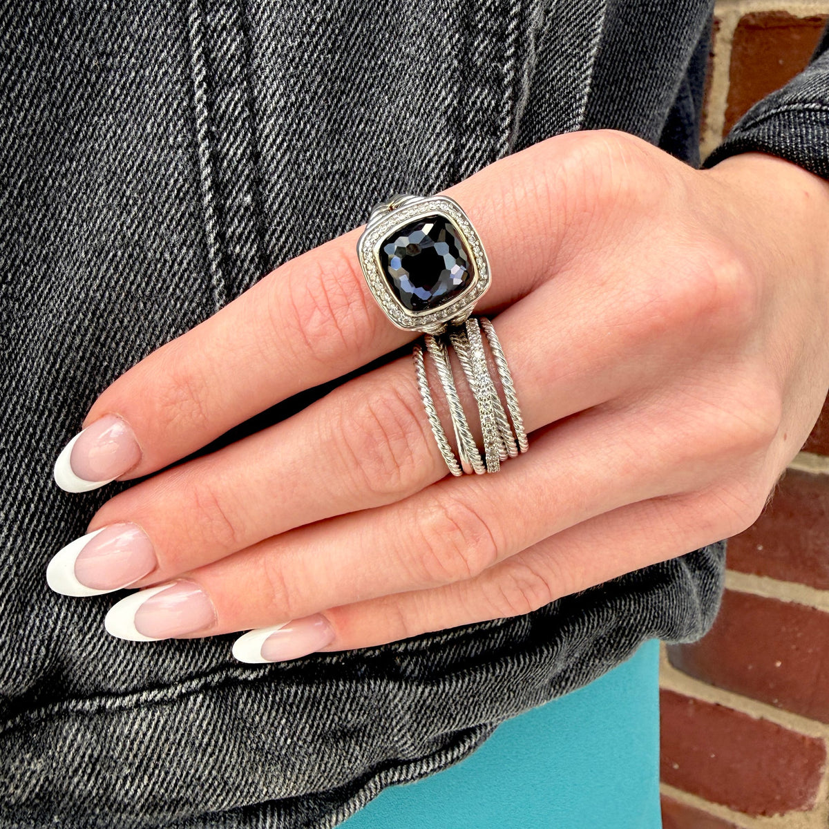 A close-up of a hand wearing a silver-toned ring with a square-cut dark blue gemstone, surrounded by a halo setting and a matching multi-band ring, against a denim jacket and brick wall background.