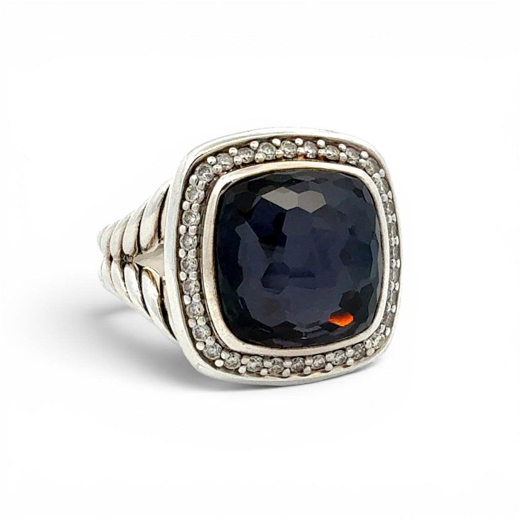 A silver-toned ring featuring a large, cushion-cut dark navy blue gemstone surrounded by a halo of small clear stones, with a textured band, set against a plain white background.