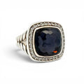 A silver-toned ring featuring a large, cushion-cut dark navy blue gemstone surrounded by a halo of small clear stones, with a textured band, set against a plain white background.