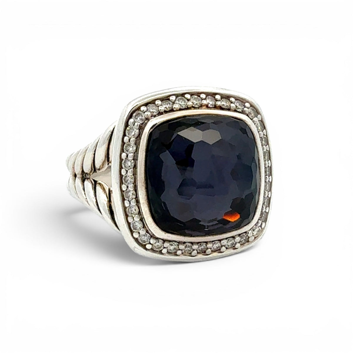 A silver-toned ring featuring a large, cushion-cut dark navy blue gemstone surrounded by a halo of small clear stones, with a textured band, set against a plain white background.