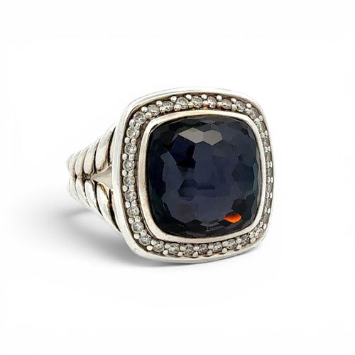 A silver-toned ring featuring a large, cushion-cut dark navy blue gemstone surrounded by a halo of small clear stones, with a textured band, set against a plain white background.