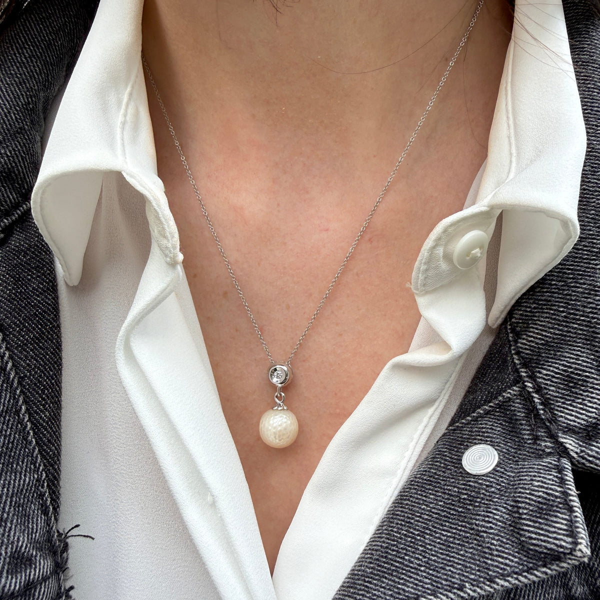 A close-up of a person wearing a delicate silver chain necklace with a textured white pearl pendant and a small clear stone accent, styled over a white collared shirt and a dark denim jacket.