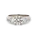 A three-stone diamond ring featuring a large round center stone flanked by two smaller round stones, set in a polished silver-colored metal band on a white background.