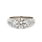 A three-stone diamond ring featuring a large round center stone flanked by two smaller round stones, set in a polished silver-colored metal band on a white background.