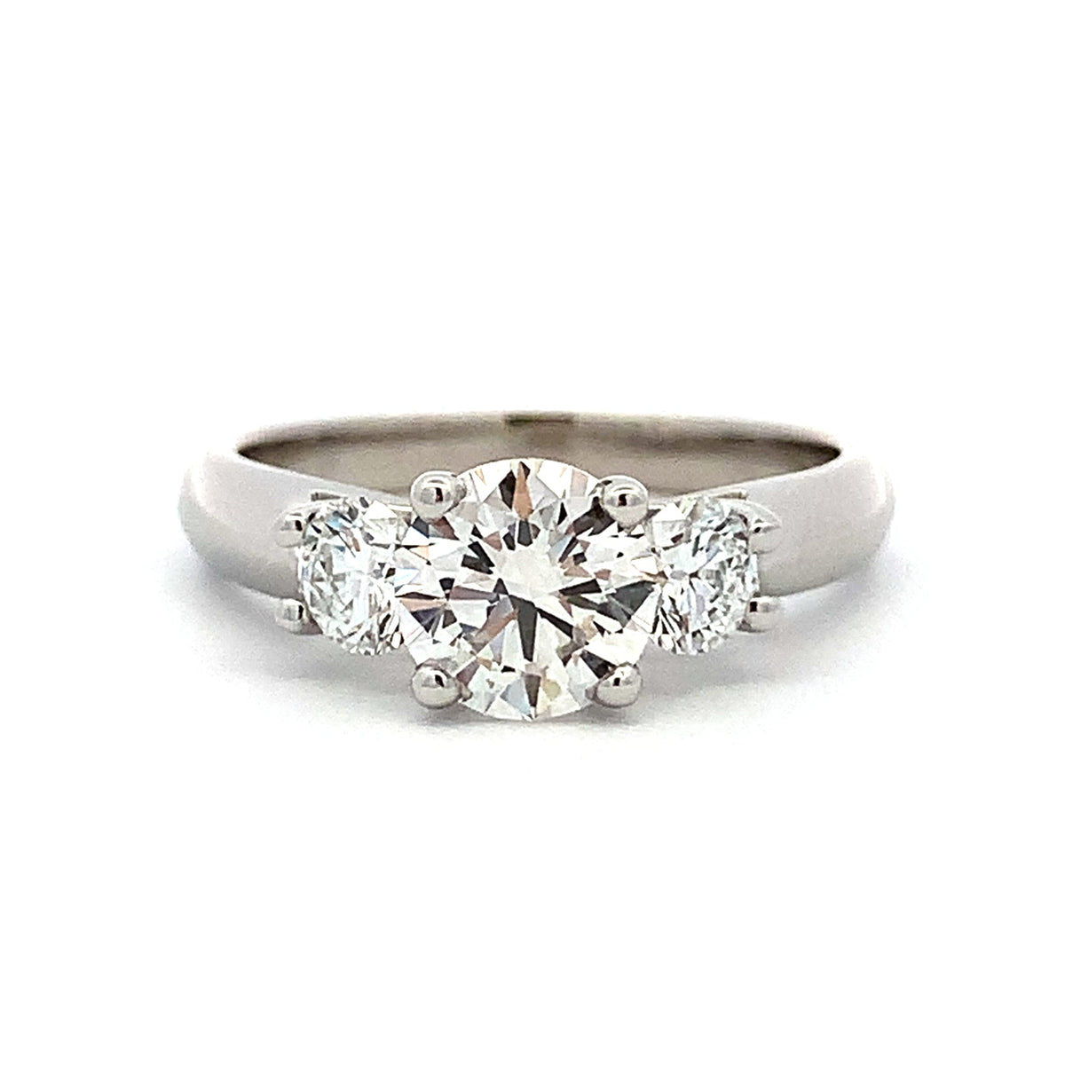 A three-stone diamond ring featuring a large round center stone flanked by two smaller round stones, set in a polished silver-colored metal band on a white background.
