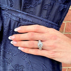 A close-up of a hand wearing a sparkling diamond engagement ring, resting on a navy blue embroidered dress against a brick wall background.