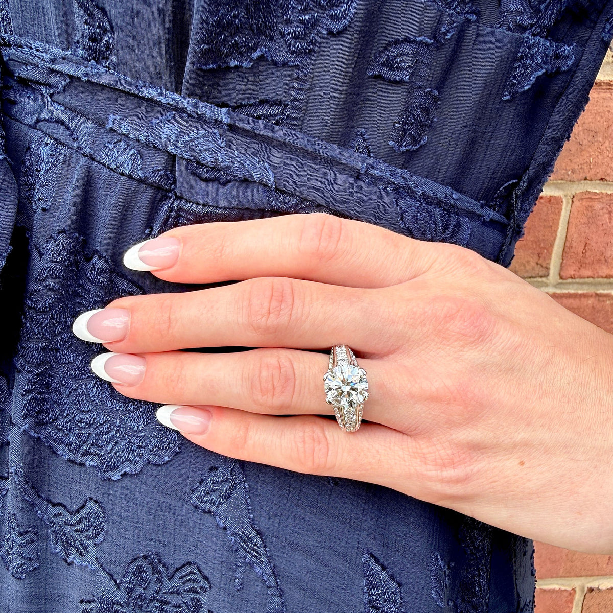 A close-up of a hand wearing a sparkling diamond engagement ring, resting on a navy blue embroidered dress against a brick wall background.