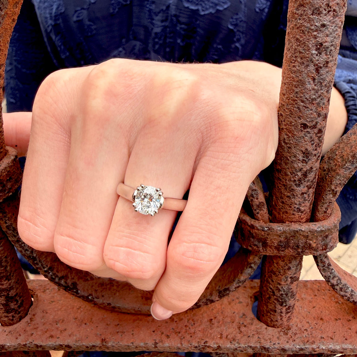 A close-up of a hand wearing a classic solitaire engagement ring with a round brilliant-cut diamond on a thin gold band, resting against a rusty metal gate.