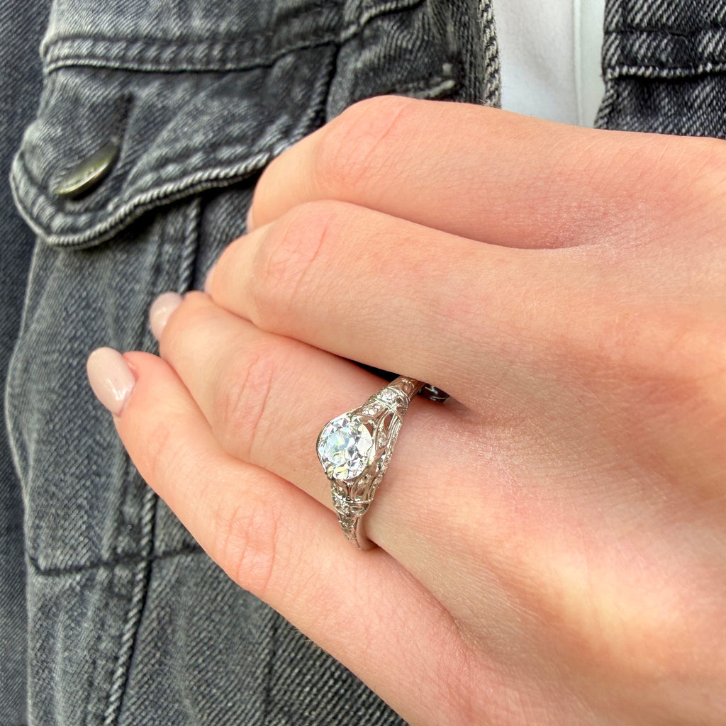A close-up of a hand wearing a silver-toned split-shank ring with a round-cut clear center stone and small side stones, shown against denim clothing.