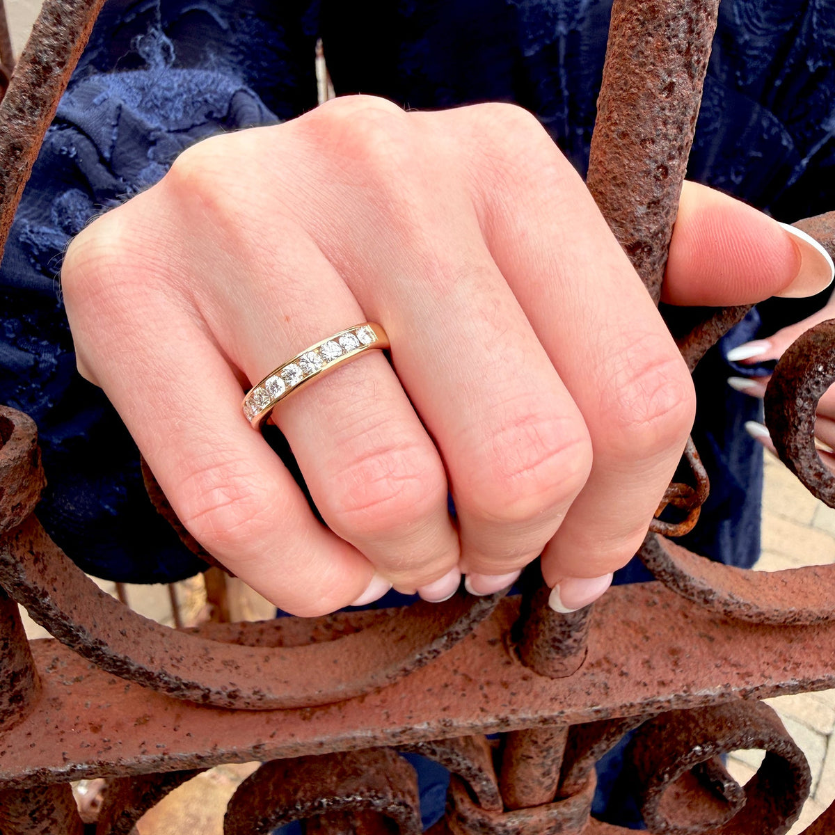 A close-up of a hand wearing a yellow gold eternity ring with small clear stones, resting on a rusty wrought iron fence.